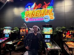 Eigenaar van het Dutch Pinball Museum, Gerard van de Sanden, staat voor vier Jurassic Park-flipperkasten en een groot neonbord in verschillende kleuren met de naam van het museum erop.