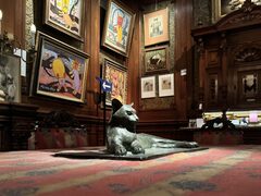 Een bronzen beeld van een liggende kat ligt op een rood beklede tafel. Op de achtergrond hangt allerlei kleurrijke schilderkunst met katten erop.