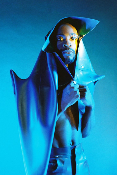 Artiest Isaï staat in blauw licht voor een blauwe achtergrond. Om zich heen draagt hij een soort sculptuur-achtige jas. Hij kijkt recht in de camera, bij zijn rechteroog draagt hij grote nepwimpers.