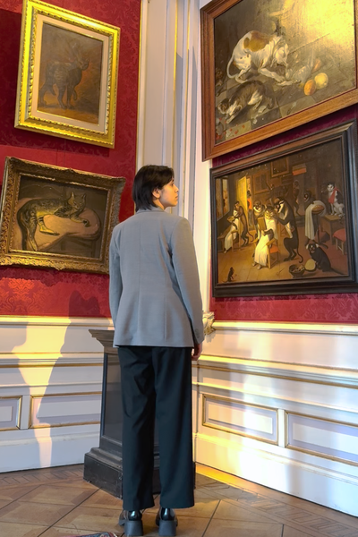 Redacteur Seshiekela staat voor een hoge muur in een Amsterdams grachtenpand. Aan de muur hangen allerlei oude schilderijen met kattenkunst eraan.