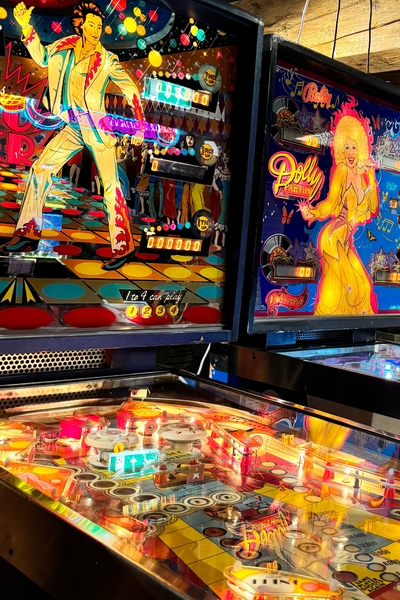 Twee lichtgevende flipperkasten naast elkaar. Links één in een discothema met een dansende man erop en rechts één met een Dolly Parton-thema.