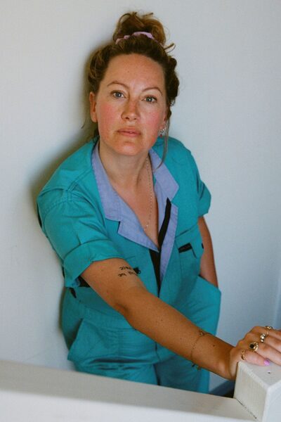 Actrice Laurien van Rijswijk staat tegen een witte muur. Ze draagt een blauwe overal en haar haar in een knot.