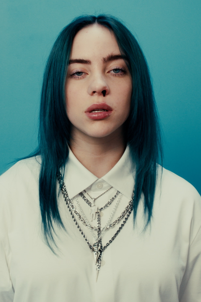 Zangeres Billie Eilish staat voor een blauwe muur. Ze heeft blauw haar, draagt een wit overhemd met kettingen erover en er komt bloed uit haar neus.