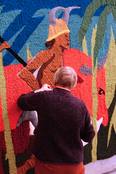 Er wordt door twee vrouwen gewerkt aan het wandtapijt. Op het tapijt staan twee personen, gebroken ketens, nootmuskaat en een palmboom.
