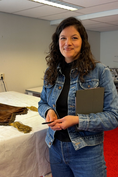 Lorena in haar atelier. Ze heeft een spijkerpak aan en los bruin haar. Ze lacht naar de camera. Ze heeft een tablet en pen in haar hand.