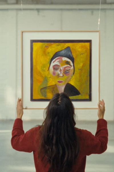 Vrouw kijkt naar een geel schilderij van een vrouw met een hoedje dat ze in haar handen heeft