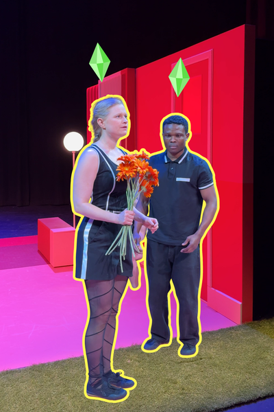 Een stil uit de voorstelling. we zien links een vrouw met oranje bloemen in haar hand en rechts een man die naar de bloemen kijkt. Ze staan stil alsof ze poppen zijn. Het decor waar ze instaan is roze en oranje.