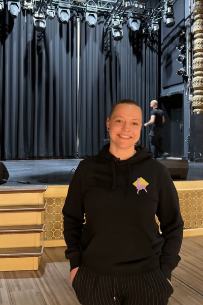 Izanka, de geïnterviewde, in de zaal. Ze heeft zwarte kleren aan en heeft haar handen in haar zakken. Ze kijkt lachend de camera in.