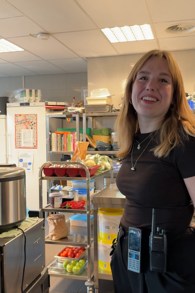 Een foto van Susanne die bij een keuken staat. Susanne is een jonge meid met los blond haar en ze kijkt lachend naar de camera. Achter haar zie je een grote professionele keuken.