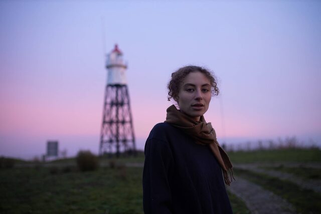 Sarah Neutkens staat bij een spoor tegen een paarse lucht aan en kijkt de camera in.
