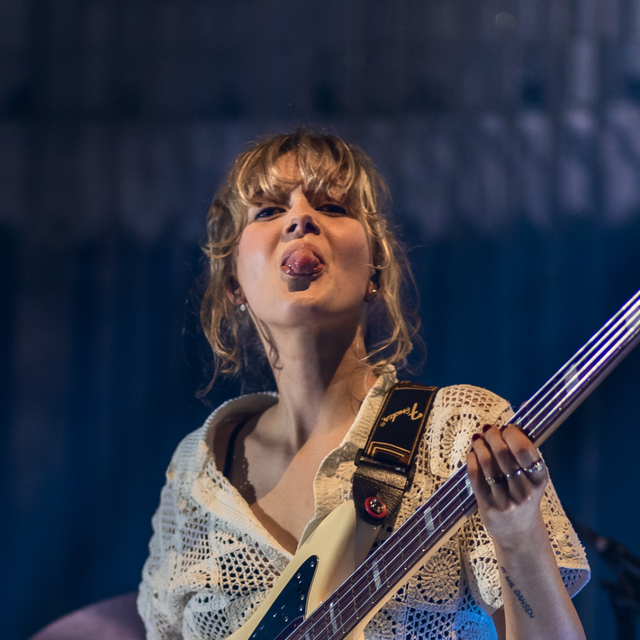 Een foto van Pieteke, een blonde meid met pony. Ze staat op een podium met een basgitaar in haar handen en steekt haar tong uit.