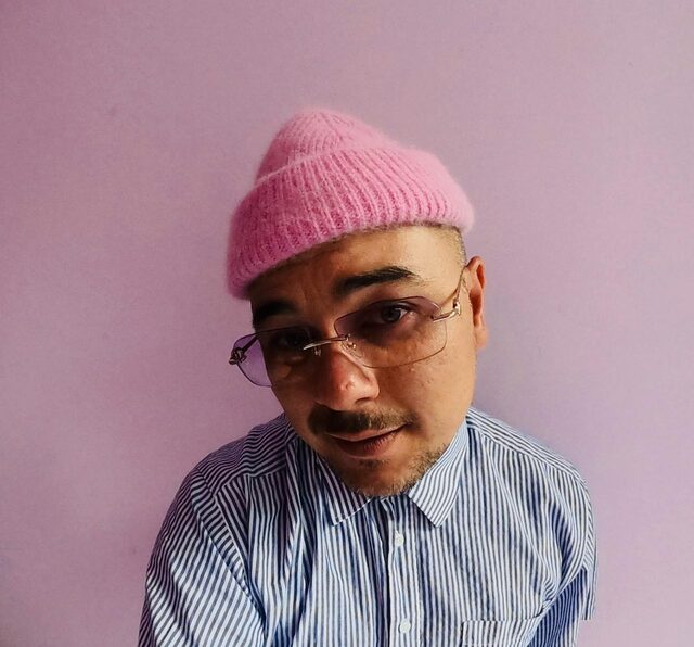 Een foto van (radio) DJ Justin Verkijk. Hij staat voor een roze achtergrond en heeft een roze muts op. Hij houdt zijn hoofd een beetje scheef en kijkt de camera in.