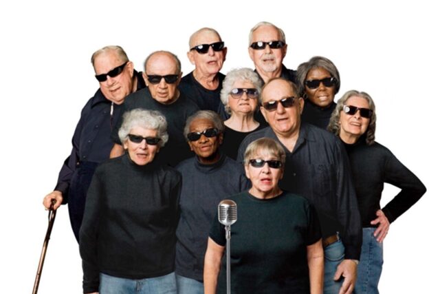 Een groep koorleden uit de documentaire zingt voor een microfoon. Ze dragen zwarte shirts en zwarte zonnebrillen.