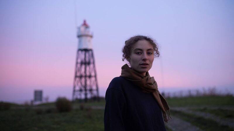 Sarah Neutkens staat bij een spoor tegen een paarse lucht aan en kijkt de camera in.