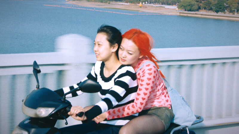 Twee Chinese actrices uit de film 1 Girl Infinite zitten samen op een scooter. Degene die rijdt, draagt een zwart/wit gestreept shirt en heeft zwart haar. Degene die achterop zit, houdt de andere persoon vast om haar middel. Ze draagt een roze shirt met rode hartjes, een groen rokje en heeft felrood haar.