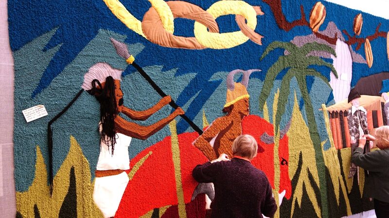 Er wordt door twee vrouwen gewerkt aan het wandtapijt. Op het tapijt staan twee personen, gebroken ketens, nootmuskaat en een palmboom.