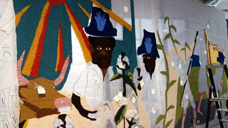 Deel van het wandkleed dat nog in constructie is. Je ziet een zon en twee mannen.