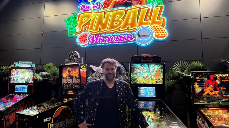 Eigenaar van het Dutch Pinball Museum, Gerard van de Sanden, staat voor vier Jurassic Park-flipperkasten en een groot neonbord in verschillende kleuren met de naam van het museum erop.