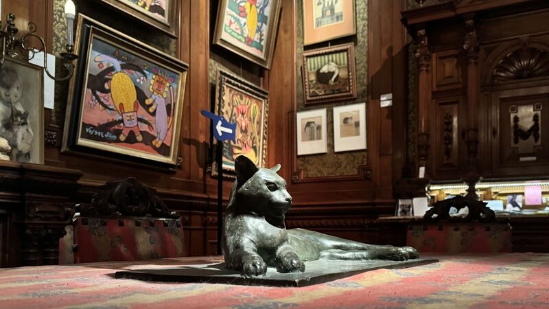 Een bronzen beeld van een liggende kat ligt op een rood beklede tafel. Op de achtergrond hangt allerlei kleurrijke schilderkunst met katten erop.