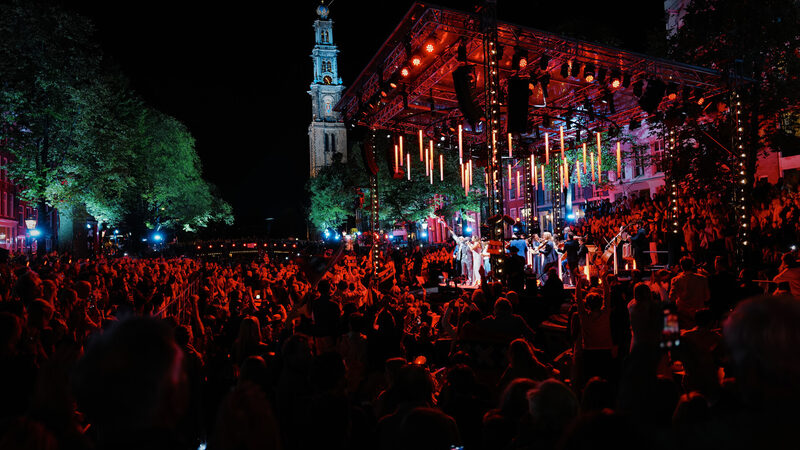 Honderden mensen staan in rood licht in het donker buiten bij de Prinsengracht in Amsterdam met een groot podium vol musici en de Westertoren op de achtergrond
