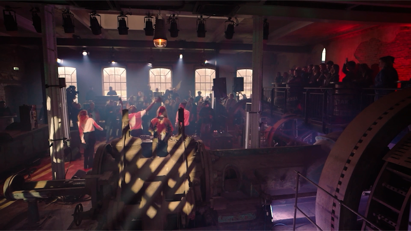 een stilbeeld van de livesessie opnames. Je ziet een grote fabriekshal waar je op de rug een band ziet spelen.