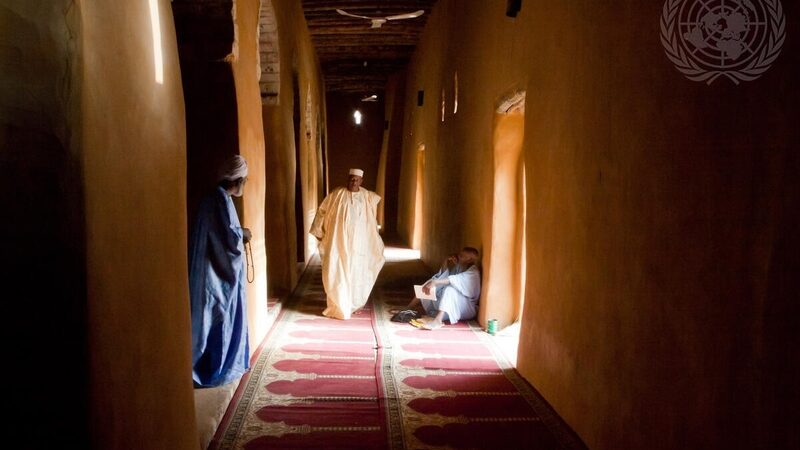 Drie mannen in een huis. Links staat een man in een blauw gewaad. In het midden loopt een man in een wit gewaad door de gang. Rechts zit een man op de grond.