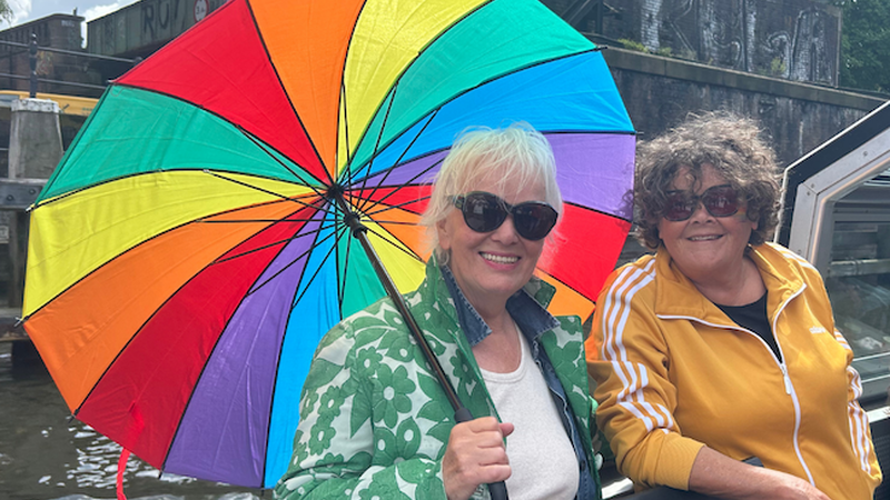 Twee dames op het dek van de boot. Links heeft een mevrouw een een regenboogparaplu vast.