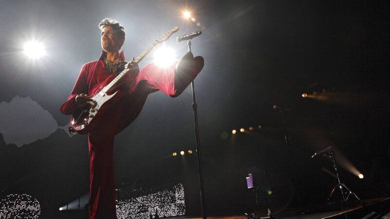 Persoon in een rood pak met een rode gitaar staat te spelen op een podium