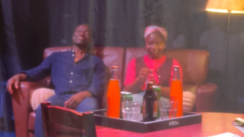 Op deze foto zien we de vader en dochter uit het theaterstuk op de bank op het toneel zitten. Er hangt een gaasdoek tussen ons en het podium in.