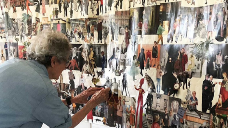Kunstenaar Aldwyth staat voorover gebogen voor haar collagewerk. Het kunstwerk bestaat uit een hele muur vol uitgeknipte afbeeldingen en illustraties.