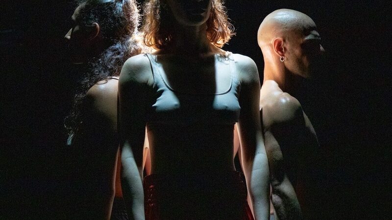 Drie dansers staan op een donker toneel.