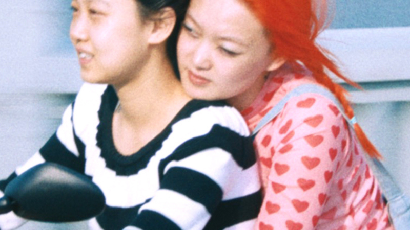 Twee Chinese actrices uit de film 1 Girl Infinite zitten samen op een scooter. Degene die rijdt, draagt een zwart/wit gestreept shirt en heeft zwart haar. Degene die achterop zit, houdt de andere persoon vast om haar middel. Ze draagt een roze shirt met rode hartjes, een groen rokje en heeft felrood haar.