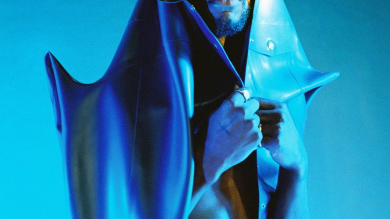 Artiest Isaï staat in blauw licht voor een blauwe achtergrond. Om zich heen draagt hij een soort sculptuur-achtige jas. Hij kijkt recht in de camera, bij zijn rechteroog draagt hij grote nepwimpers.