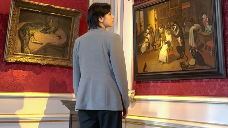 Redacteur Seshiekela staat voor een hoge muur in een Amsterdams grachtenpand. Aan de muur hangen allerlei oude schilderijen met kattenkunst eraan.