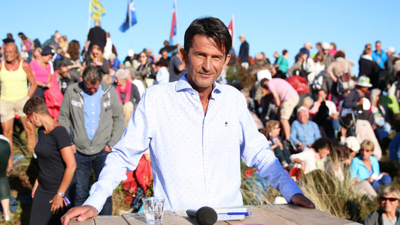 Cornald Maas staat glimlachend in een overhemd achter zijn tafel op de set van Opium op Oerol op Terschelling met heel veel mensen achter hem
