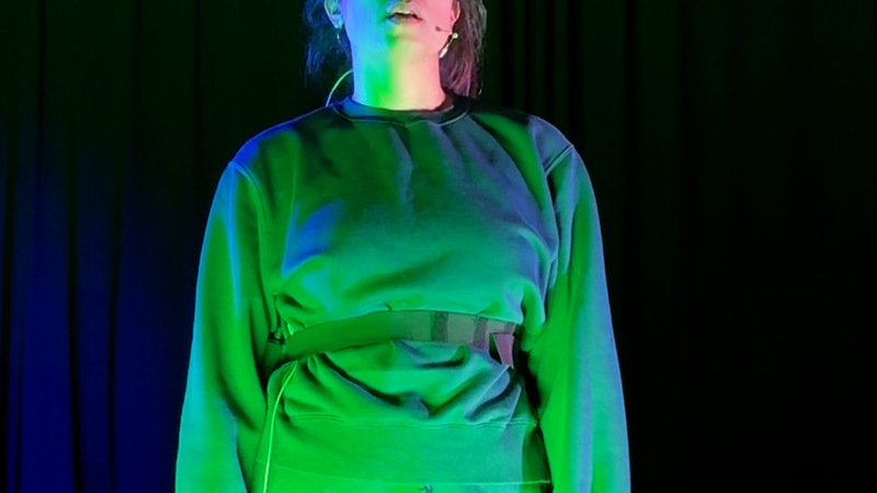 Foto van Yeliz op het toneel. Ze heeft een blauw pak aan en is groen uitgelicht. Ze kijkt serieus.