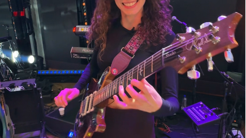 We zien Yvette met haar gitaar in haar handen. Ze staat in een tv studio en kijkt de camera in en lacht. Ze speelt op haar gitaar.