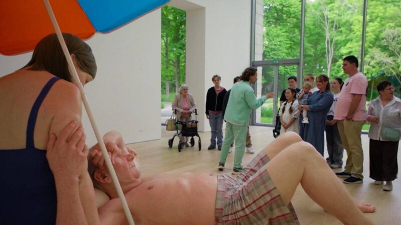 Een groep bezoekers kijkt in Museum Voorlinden naar een kunstwerk van twee levensgrote figuren in badkleding onder een strandparasol. Op de achtergrond geeft een gids uitleg in de lichte museumzaal.