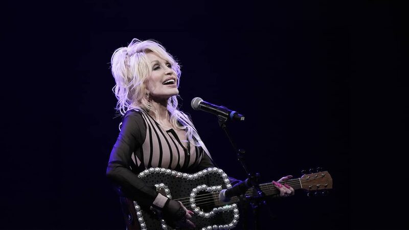 Dolly Parton staat zingend op een podium met een gitaar waarvan de rand is versierd met grote parels.