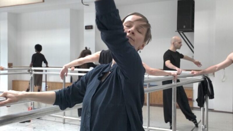 Een foto van danseres Jozefien Debaillie. Ze staat voor een spiegel aan de bar. Ze heeft haar ogen dicht en een arm omhoog. In de achtergrond zie je via de spiegel ook andere balletdansers in de zaal aan het oefenen.