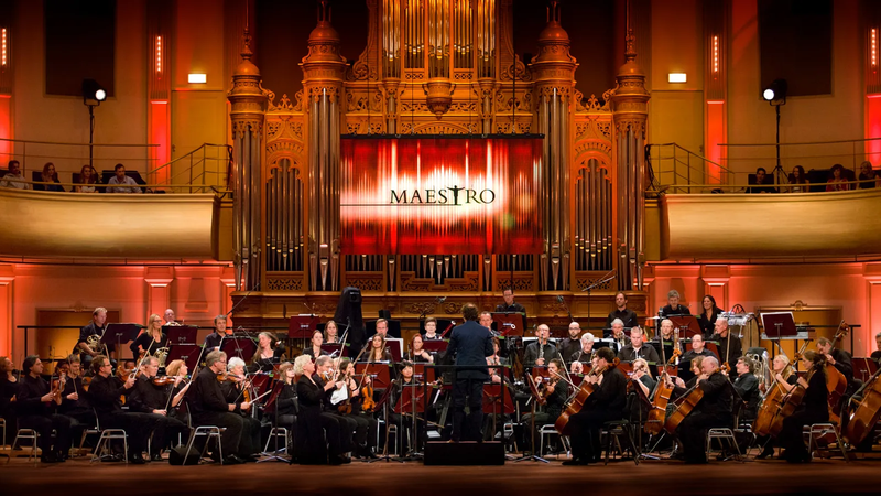 Groot klassiek orkest zit op het podium te spelen met maestro groot op het scherm achter het orkest