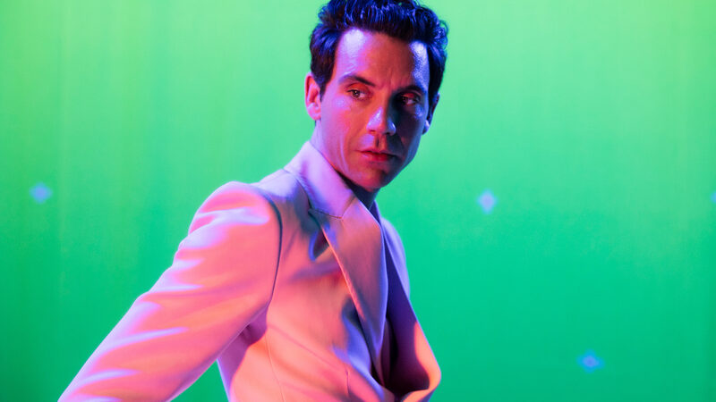 Foto van zanger Mika, in een roze pak voor een groene achtergrond.