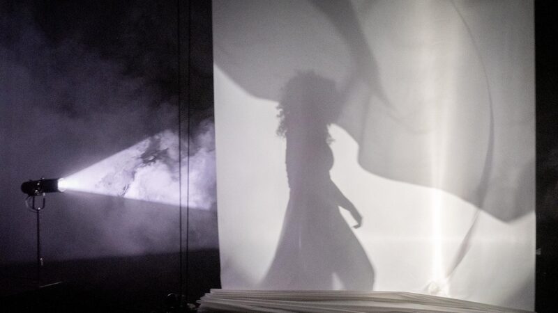 Silhouet van een dansende vrouw achter een wit doek omring met lichten in een theaterzaal