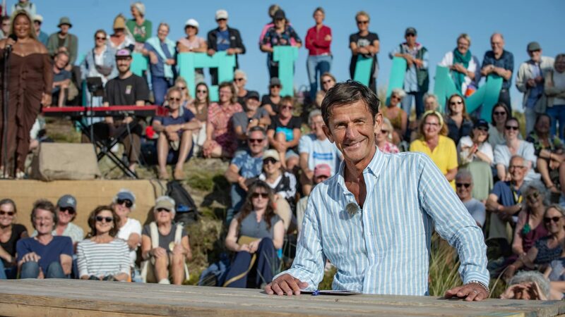 Cornald Maas staat glimlachend in een overhemd achter zijn tafel op de set van Opium op Oerol op Terschelling met heel veel mensen achter hem