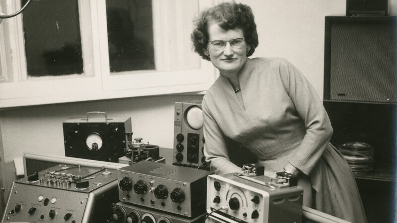 Componist op zwart wit foto omringd door apparatuur, waarop ze elektronische muziek componeert.
