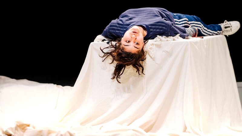 Actrice Soumaya ligt in blauwe kleding met haar rug op een tafel met een groot wit doek. Haar hoofd hangt over de tafel heen en ze kijkt naar rechts.