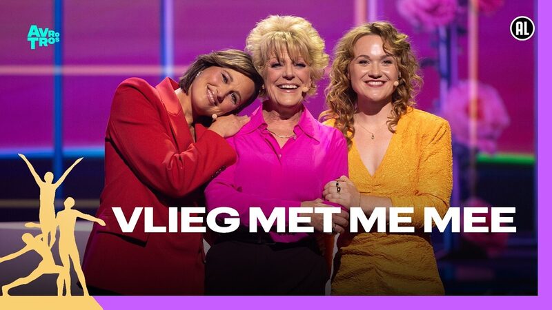 Drie vrouwen in gekleurde kleding staan glimlachen naast elkaar op een podium met een roze achtergrond