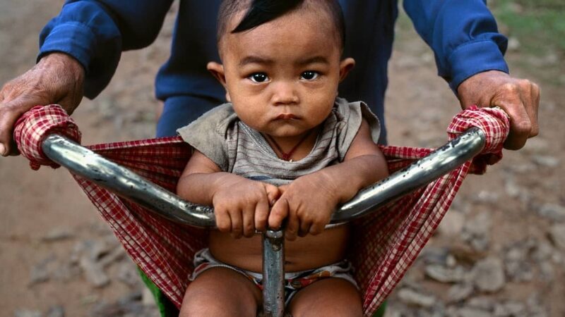 Baby in a sling at Banteay Srei, Angkor, Cambodia, 2000
Een klein Cambodjaans jongetje zit voorop de fiets bij iemand. Hij zit in een rode geknoopte doek die aan het stuur hangt. Het kleine jongetje kijkt recht de camera in. Hij heeft een donkere lok haar, een blote buik en blote benen. Zijn kleren zien er stoffig uit.