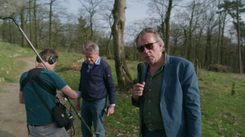 Man met een zonnebril staat met een microfoon in zijn hand in de natuur