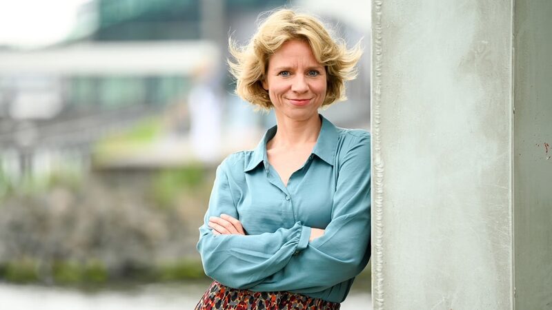 Portretfoto van Floortje Smit op een stedelijke achtergrond. Ze draagt een blauwe blouse met haar armen over elkaar.